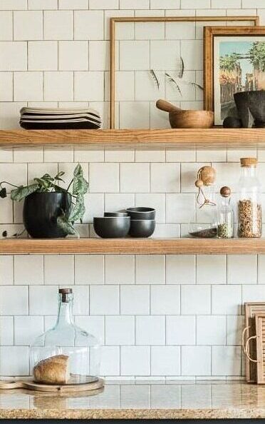 Minimalist kitchen shelves adorned with black bowls, a black plant pot, a round glass jar with cork lid, stacked dishes, and framed artwork. The shelves front a white tiled wall and a counter with a large glass decanter and a loaf of bread on a wooden board.