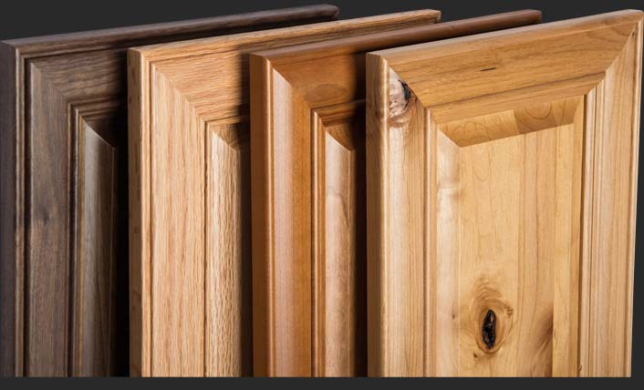 A selection of wooden cabinet doors in various shades and types of wood finishes, displayed vertically side by side. The finishes include dark brown, light wood, medium brown, and a blend of natural wood grains, each with a raised panel design.