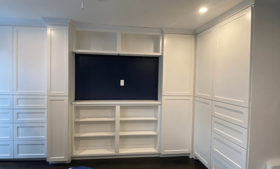 A built-in shelving and cabinet unit is shown, featuring a combination of white cabinets and open shelves with a central recessed area painted dark blue. The unit has tall cabinets on either side and additional cabinets surrounding the open shelving.