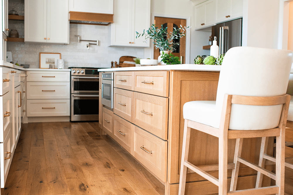 A modern kitchen featuring a wooden island with storage drawers and a white upholstered bar stool. The island is topped with a plant and various vegetables. White cabinets and stainless steel appliances are visible, with hardwood floors completing the look.