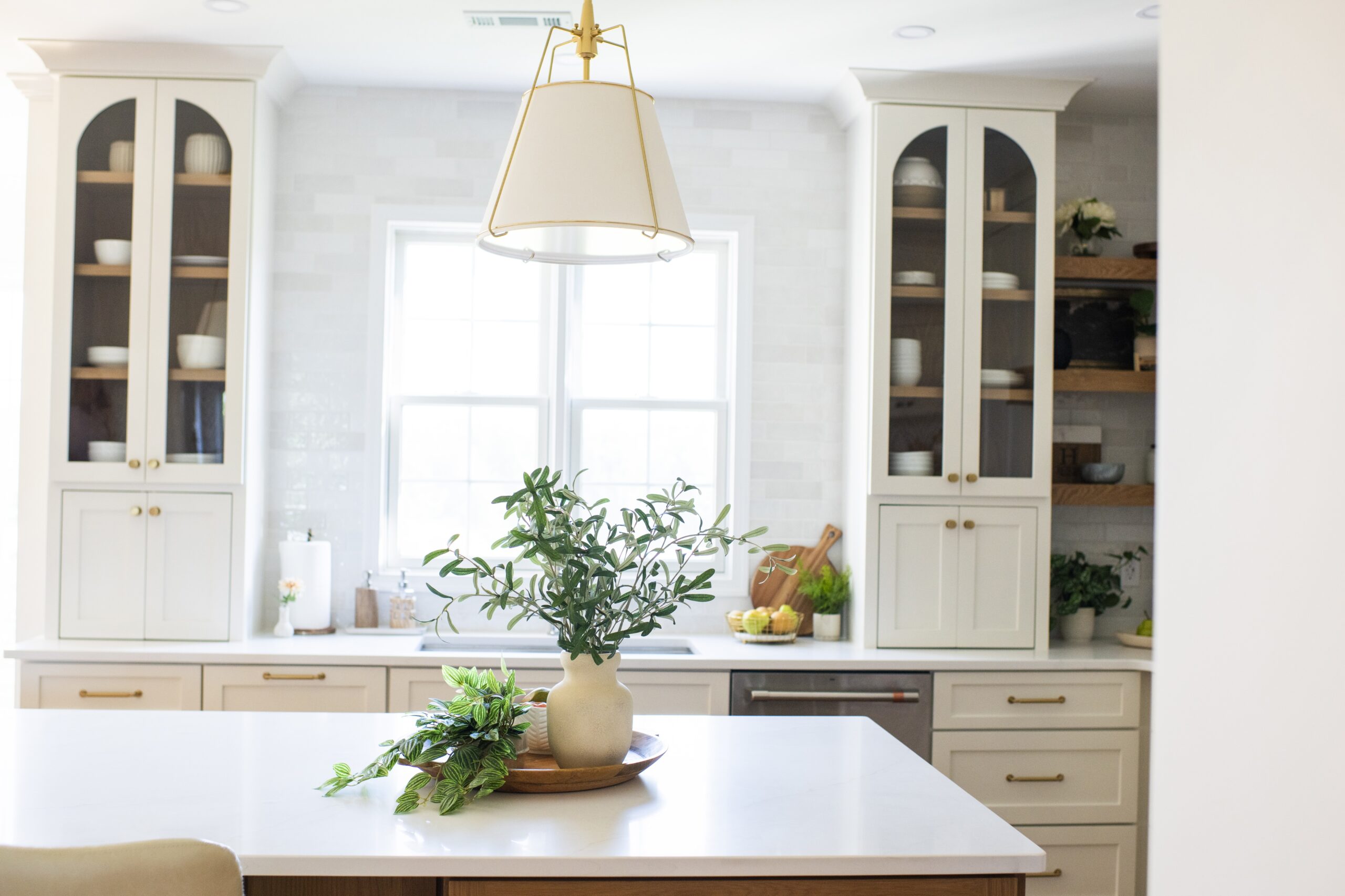 A bright, modern kitchen with white cabinets, glass-paneled upper doors, brass hardware, and open wooden shelves. A large window above the counter illuminates the space. A white island with a plant centerpiece is in the foreground, and a stylish pendant light hangs above it.