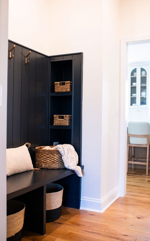 A modern mudroom featuring a dark blue paneled wall with hooks and shelves holding wicker baskets. The bench below has a large throw pillow and a plaid blanket draped over a basket. The space has wooden flooring and leads into a brightly lit kitchen area.